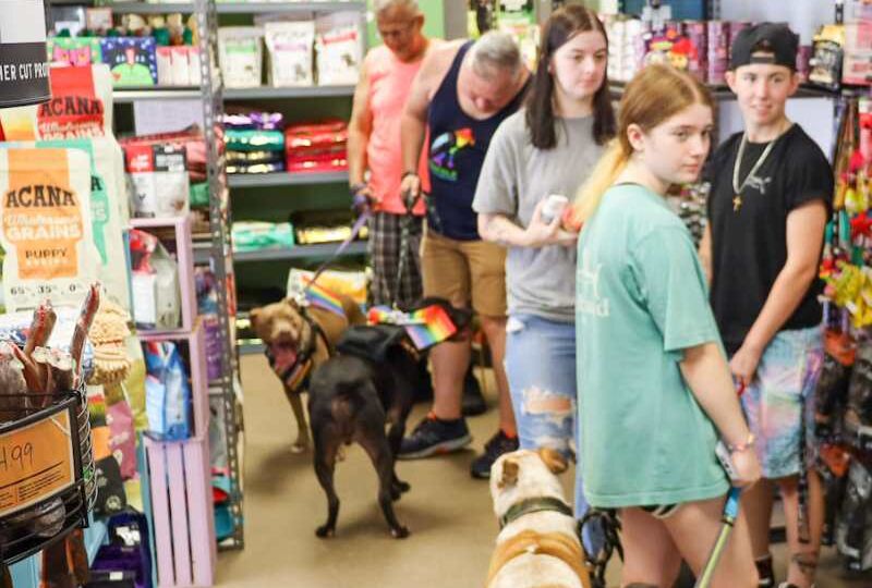 A lively in-store retail event with shoppers browsing, engaging with products, and a store owner welcoming customers, creating a strong community vibe.